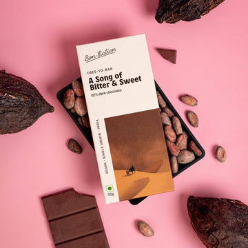 A Song of Bitter and Sweet - 82% Dark on a bed of cocoa beans, on a light pink table with cacao pods and unwrapped chocolate from Bon Fiction