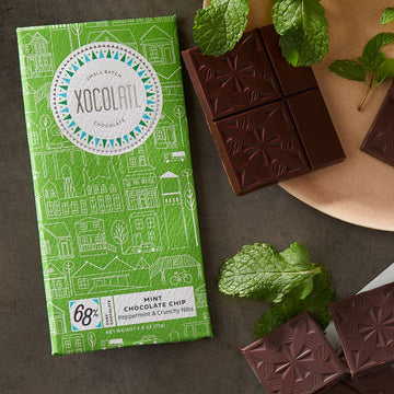 Mint Chocolate Chip 68% Dark on a grey table with broken pieces of unwrapped chocolate, and peppermint leaves