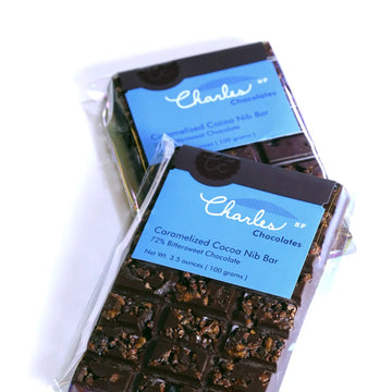 Two wrapped Caramelized Cocoa Nib Bars from Charles Chocolates in clear packaging, laying on top of one another, against a white background.