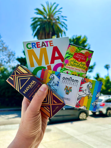 Hand holds four chocolate bars against a sunny sky with palm trees to celebrate pride month