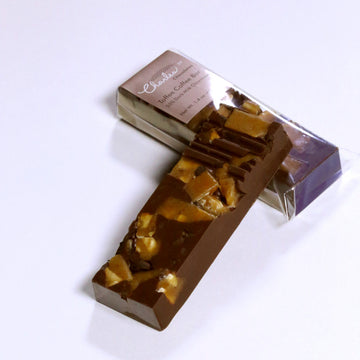 An unwrapped Toffee Coffee bar laying on top of a wrapped Toffee Coffee bar, against a white background.