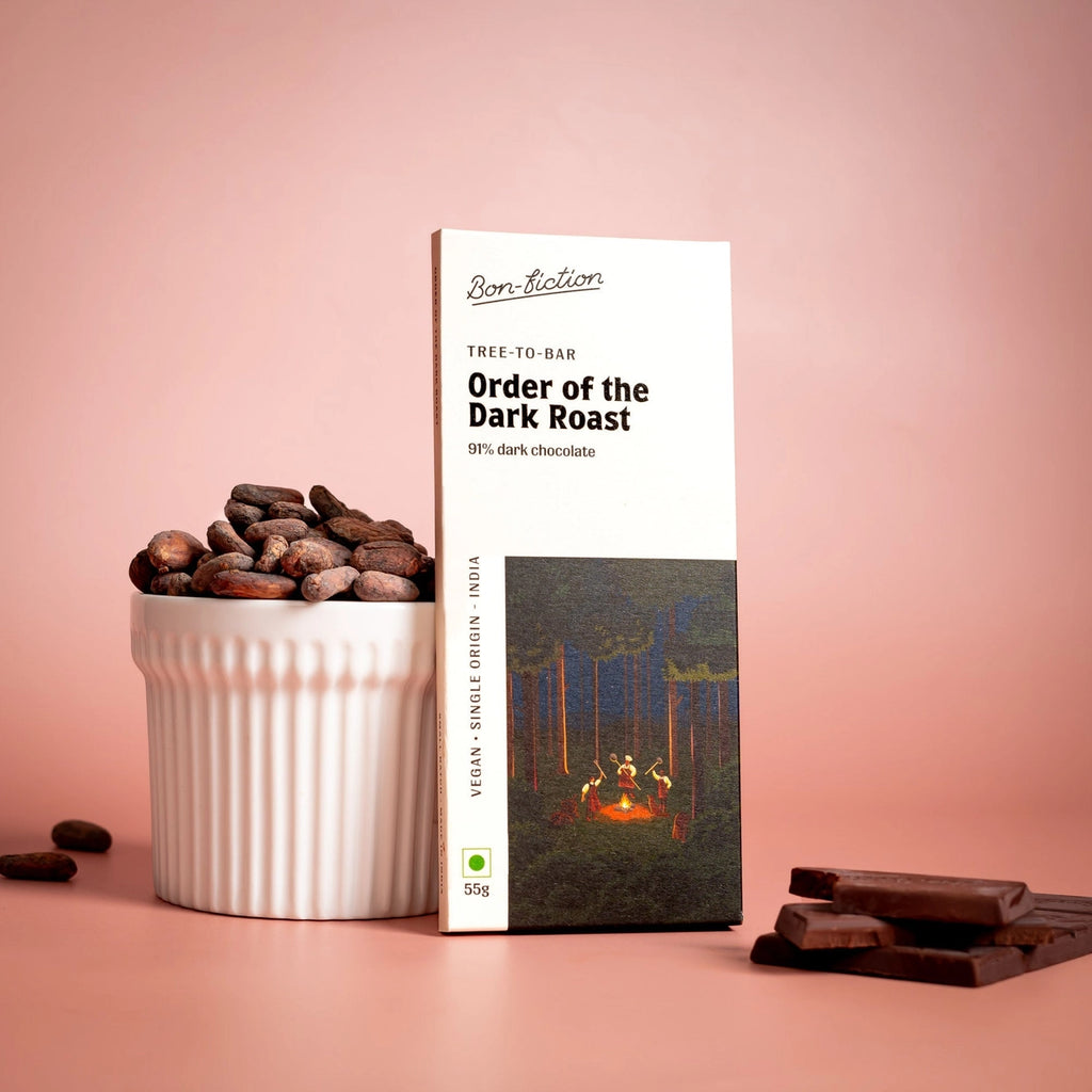 The front of a wrapped "Order of the Dark Roast" chocolate bar standing upright against a cup of cocoa beans, with pieces of the unwrapped "Order of the Dark Roast" chocolate bar to the right of it. There is a tinted background.