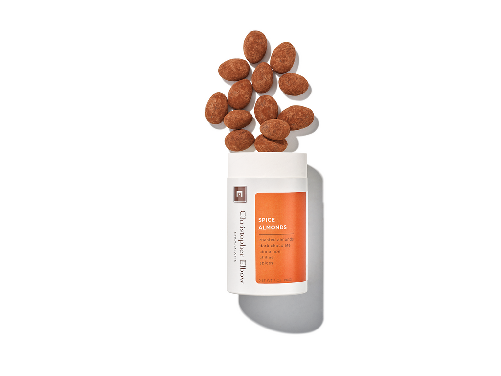 A jar of Spiced Dark Chocolate Almonds from Christopher Elbow, with a few Spiced Dark Chocolate Almonds above the jar, against a white background.