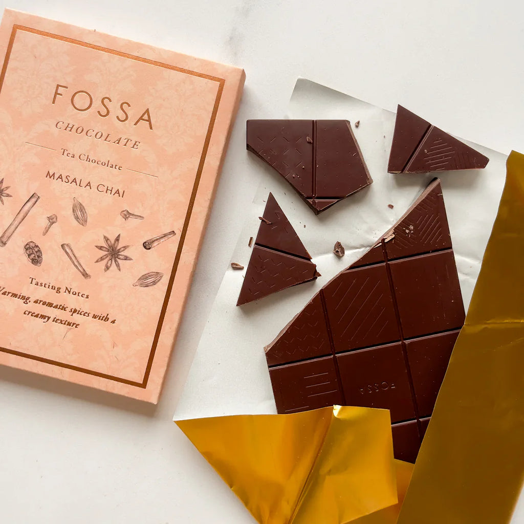 The front of Fossa's wrapped Masala Chai Chocolate bar, with a broken unwrapped Masala Chai Chocolate Bar next to it with partial gold wrapping on the bottom.